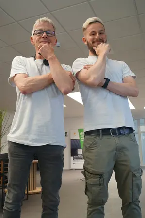 Zwei Männer stehen im modernen Büro, beide tragen helle T-Shirts und wirken nachdenklich.