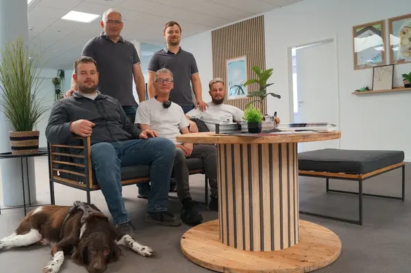 Fünf Männer und ein Hund sitzen entspannt im modernen Büro mit Pflanzen und Holztisch.